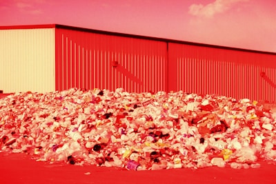 A large pile of assorted trash, including plastic bottles, bags, and various discarded items, is seen in front of an industrial building. The entire scene is bathed in a red hue, likely due to the lighting or color editing.