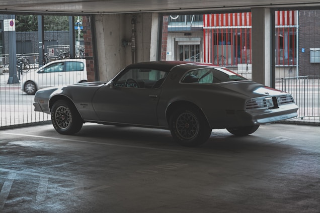 A vintage car is parked inside a dimly lit garage. The car, likely a classic model, features sleek lines and a silver paint finish. Its surroundings include concrete walls and a striped red-and-white exterior visible through the open sides of the garage.