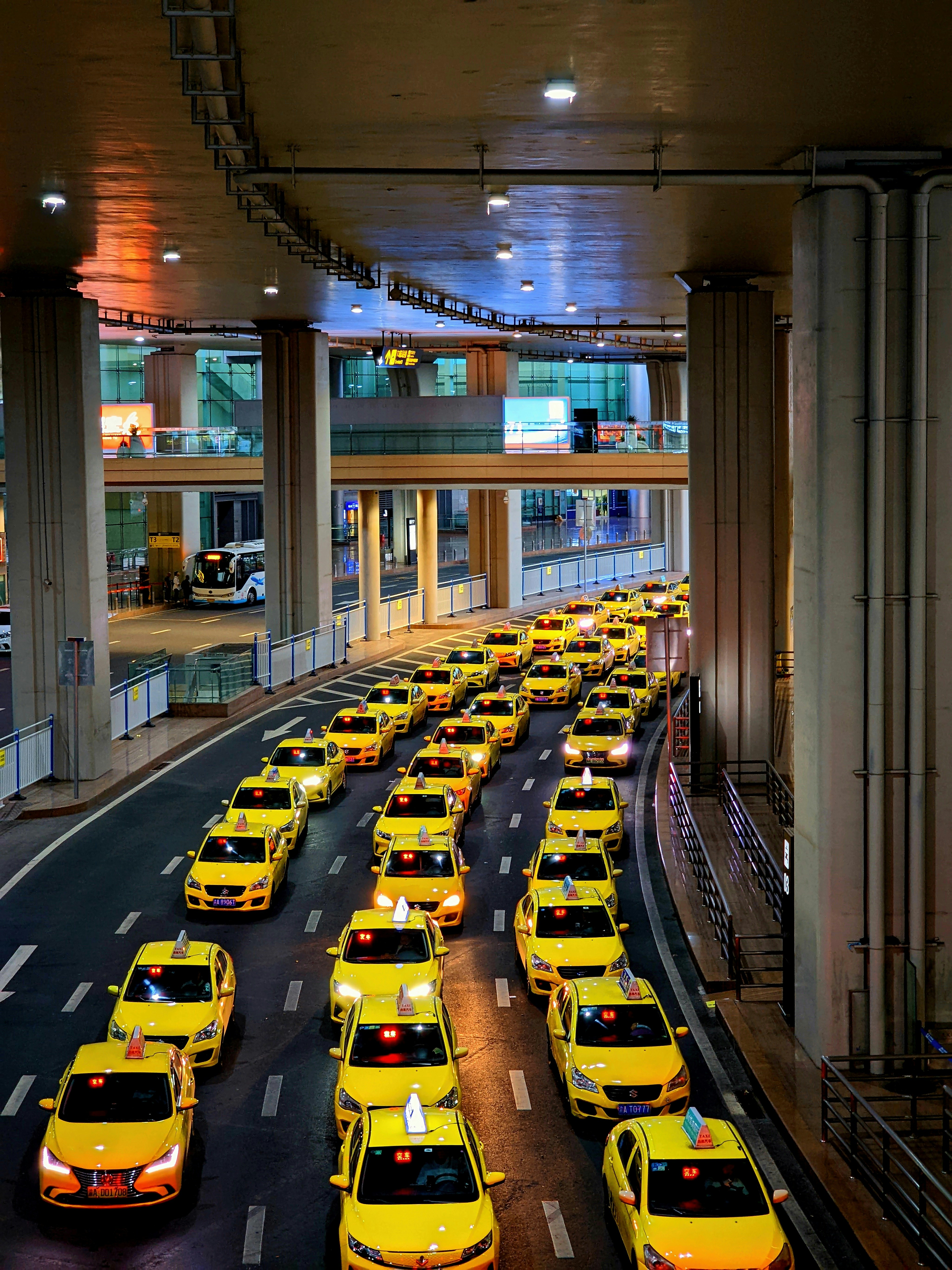 A long line of bright yellow taxis waits in orderly rows beneath a high, industrial ceiling. The setting appears to be a modern transportation hub, with multiple levels observed in the background. The scene is well-lit, creating a vibrant and bustling atmosphere.