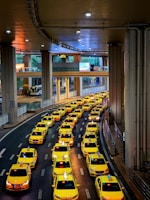A long line of bright yellow taxis waits in orderly rows beneath a high, industrial ceiling. The setting appears to be a modern transportation hub, with multiple levels observed in the background. The scene is well-lit, creating a vibrant and bustling atmosphere.
