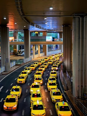 A long line of bright yellow taxis waits in orderly rows beneath a high, industrial ceiling. The setting appears to be a modern transportation hub, with multiple levels observed in the background. The scene is well-lit, creating a vibrant and bustling atmosphere.