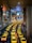 A long line of bright yellow taxis waits in orderly rows beneath a high, industrial ceiling. The setting appears to be a modern transportation hub, with multiple levels observed in the background. The scene is well-lit, creating a vibrant and bustling atmosphere.