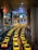 A long line of bright yellow taxis waits in orderly rows beneath a high, industrial ceiling. The setting appears to be a modern transportation hub, with multiple levels observed in the background. The scene is well-lit, creating a vibrant and bustling atmosphere.