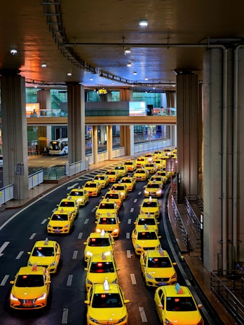 A long line of bright yellow taxis waits in orderly rows beneath a high, industrial ceiling. The setting appears to be a modern transportation hub, with multiple levels observed in the background. The scene is well-lit, creating a vibrant and bustling atmosphere.
