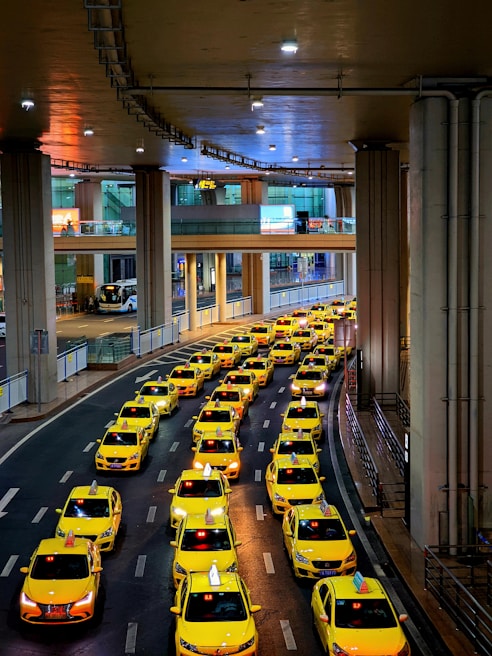 A long line of bright yellow taxis waits in orderly rows beneath a high, industrial ceiling. The setting appears to be a modern transportation hub, with multiple levels observed in the background. The scene is well-lit, creating a vibrant and bustling atmosphere.