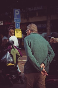 An elderly man wearing a green sweater is standing with his back to the camera, hands clasped behind him, observing the surroundings. Nearby, a person on a mobility scooter is visible, carrying a bag with fabric or clothing. A woman with a ponytail walks in the opposite direction. In the background, there are parking signs and urban elements like buildings and trees.