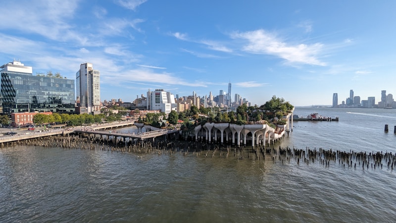 A modern cityscape featuring a waterfront with a unique park structure built on pillars extending over the water. The park is lush with vegetation and people are visible walking along the pathways. High-rise buildings and skyscrapers form the background, and the water reflects the blue sky.