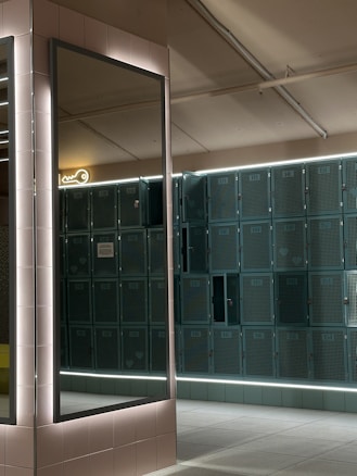 A row of teal lockers lines the wall, illuminated by overhead lights. A large, rectangular mirror framed with light-colored tiles reflects part of the room. A neon key sign is visible above the lockers, and the floor is covered with light gray tiles.
