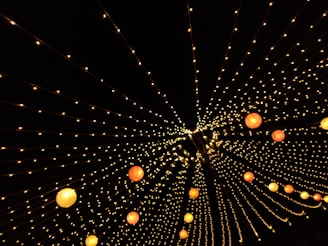 A dazzling array of string lights arranged in converging lines creates a starry effect against a dark backdrop. Round, colorful lanterns interspersed among the lights add splashes of warm color, enhancing the festive atmosphere.