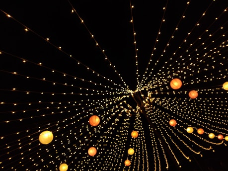 A dazzling array of string lights arranged in converging lines creates a starry effect against a dark backdrop. Round, colorful lanterns interspersed among the lights add splashes of warm color, enhancing the festive atmosphere.