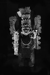 An ornate statue features intricate carvings and adornments, including a headdress with elaborate patterns, a necklace of large beads, and detailed facial expressions. Made of stone, the sculpture captures the essence of ancient symbolism and craftsmanship.