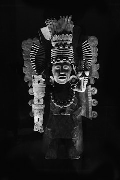 An ornate statue features intricate carvings and adornments, including a headdress with elaborate patterns, a necklace of large beads, and detailed facial expressions. Made of stone, the sculpture captures the essence of ancient symbolism and craftsmanship.