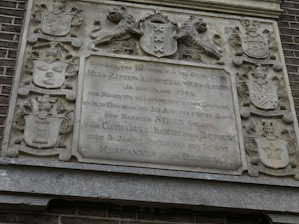 A detailed stone plaque with intricate carvings of coats of arms and a lion holding a shield. The plaque bears text in Dutch with a historical appearance, suggesting it dates back to the 18th century. The stone is weathered, indicating age, and is surrounded by brickwork.