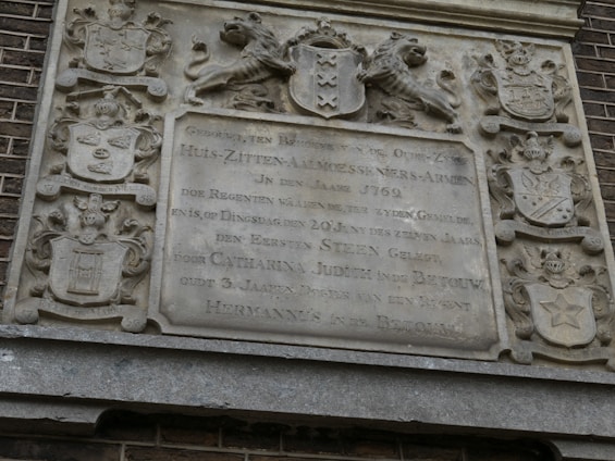 A detailed stone plaque with intricate carvings of coats of arms and a lion holding a shield. The plaque bears text in Dutch with a historical appearance, suggesting it dates back to the 18th century. The stone is weathered, indicating age, and is surrounded by brickwork.
