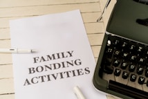 A sheet of paper with the words 'FAMILY BONDING ACTIVITIES' printed in bold, placed on a white wooden surface. There is a white pen next to the paper and a black typewriter partially visible on the right side.