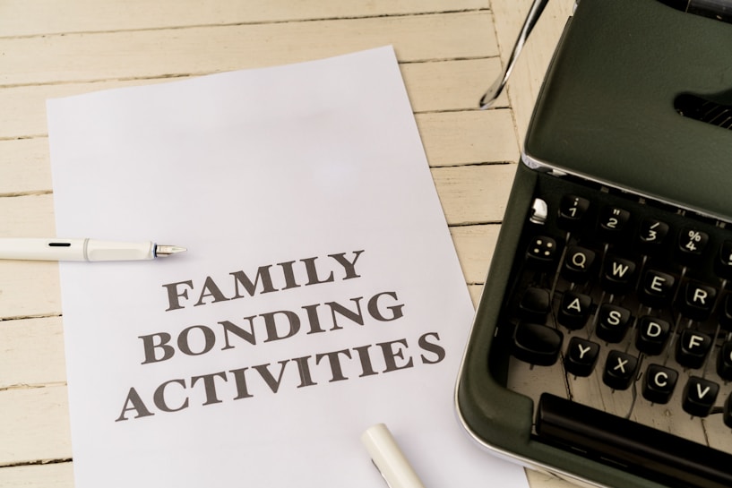 A sheet of paper with the words 'FAMILY BONDING ACTIVITIES' printed in bold, placed on a white wooden surface. There is a white pen next to the paper and a black typewriter partially visible on the right side.