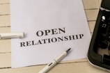 A white piece of paper laid on a wooden surface with the words 'OPEN RELATIONSHIP' printed on it. A fountain pen with a silver and black design is placed near the paper. Part of a typewriter is visible on the right side.