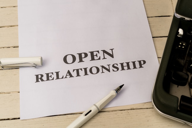 A white piece of paper laid on a wooden surface with the words 'OPEN RELATIONSHIP' printed on it. A fountain pen with a silver and black design is placed near the paper. Part of a typewriter is visible on the right side.