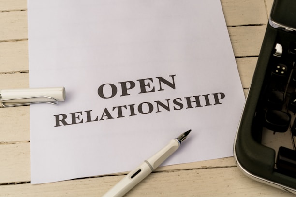 A white piece of paper laid on a wooden surface with the words 'OPEN RELATIONSHIP' printed on it. A fountain pen with a silver and black design is placed near the paper. Part of a typewriter is visible on the right side.