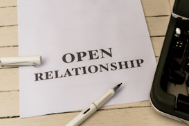 A white piece of paper laid on a wooden surface with the words 'OPEN RELATIONSHIP' printed on it. A fountain pen with a silver and black design is placed near the paper. Part of a typewriter is visible on the right side.