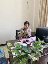 A person is sitting at a wooden desk in an office environment, engaged in a phone conversation. There is a computer monitor on the desk, along with a keyboard, a mouse, and some office documents. Several green houseplants in vases are arranged on the desk. The background features a plain wall with a power outlet and beige curtains.