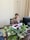 A person is sitting at a wooden desk in an office environment, engaged in a phone conversation. There is a computer monitor on the desk, along with a keyboard, a mouse, and some office documents. Several green houseplants in vases are arranged on the desk. The background features a plain wall with a power outlet and beige curtains.