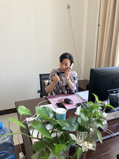 A person is sitting at a wooden desk in an office environment, engaged in a phone conversation. There is a computer monitor on the desk, along with a keyboard, a mouse, and some office documents. Several green houseplants in vases are arranged on the desk. The background features a plain wall with a power outlet and beige curtains.