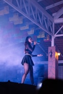 A performer on stage is holding a microphone and singing. She is wearing a black outfit with a skirt and has long hair. The stage is filled with smoke, creating a dramatic effect under purple and blue lighting. Decorative paper banners hang from the ceiling beams, adding a festive touch to the scene.