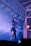 A performer on stage is holding a microphone and singing. She is wearing a black outfit with a skirt and has long hair. The stage is filled with smoke, creating a dramatic effect under purple and blue lighting. Decorative paper banners hang from the ceiling beams, adding a festive touch to the scene.