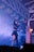 A performer on stage is holding a microphone and singing. She is wearing a black outfit with a skirt and has long hair. The stage is filled with smoke, creating a dramatic effect under purple and blue lighting. Decorative paper banners hang from the ceiling beams, adding a festive touch to the scene.