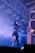 A performer on stage is holding a microphone and singing. She is wearing a black outfit with a skirt and has long hair. The stage is filled with smoke, creating a dramatic effect under purple and blue lighting. Decorative paper banners hang from the ceiling beams, adding a festive touch to the scene.