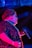 A performer is illuminated with vibrant lighting, featuring shades of blue and pink. The individual wears a sparkling sequined outfit and holds a microphone, suggesting a live performance or event. Their hair is styled neatly and they accessorize with a pearl necklace and earrings.