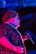 A performer is illuminated with vibrant lighting, featuring shades of blue and pink. The individual wears a sparkling sequined outfit and holds a microphone, suggesting a live performance or event. Their hair is styled neatly and they accessorize with a pearl necklace and earrings.