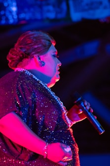 A performer is illuminated with vibrant lighting, featuring shades of blue and pink. The individual wears a sparkling sequined outfit and holds a microphone, suggesting a live performance or event. Their hair is styled neatly and they accessorize with a pearl necklace and earrings.