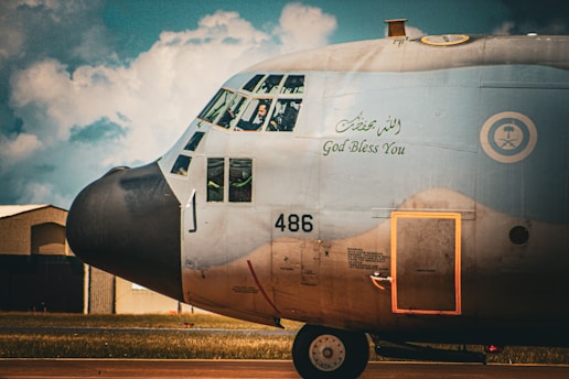 The image depicts the side view of a military aircraft with Arabic and English writing on its body. The cockpit windows reveal several pilots inside. The airplane is on a runway with a hangar and some clouds visible in the background.
