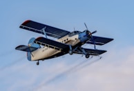 A vintage biplane in flight with a light beige fuselage and dark blue wings. The aircraft's registration is visible on the body, and it is flying against a clear blue sky.