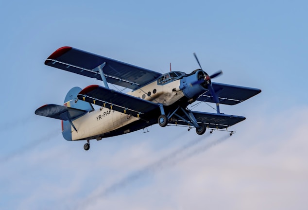 A vintage biplane in flight with a light beige fuselage and dark blue wings. The aircraft's registration is visible on the body, and it is flying against a clear blue sky.