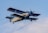 A vintage biplane in flight with a light beige fuselage and dark blue wings. The aircraft's registration is visible on the body, and it is flying against a clear blue sky.