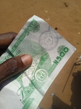 A hand is holding a 500 Naira banknote from Nigeria. The note features green and white colors with intricate designs and the denomination clearly printed. The background appears to be an outdoor sandy area.