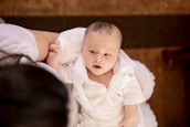 A baby wearing a white outfit lies on a soft white cushion, looking up with an expression of curiosity. The baby's skin is fair, and the clothing features a ruffled design. An adult arm, dressed in white, gently supports the child.