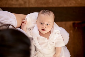 A baby wearing a white outfit lies on a soft white cushion, looking up with an expression of curiosity. The baby's skin is fair, and the clothing features a ruffled design. An adult arm, dressed in white, gently supports the child.