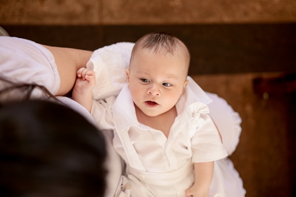 A baby wearing a white outfit lies on a soft white cushion, looking up with an expression of curiosity. The baby's skin is fair, and the clothing features a ruffled design. An adult arm, dressed in white, gently supports the child.