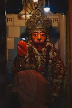 A richly adorned statue with a detailed, ornate headdress, intricate jewelry and a colorful garland. An individual dressed in traditional attire is bent forward, possibly in prayer or reverence.