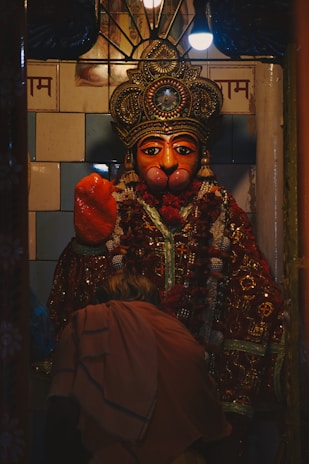A richly adorned statue with a detailed, ornate headdress, intricate jewelry and a colorful garland. An individual dressed in traditional attire is bent forward, possibly in prayer or reverence.