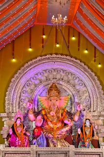 An elaborate display featuring a large, colorful statue of a deity with an elephant head, adorned with vibrant garments and surrounded by intricate silver decorations. Flanking the central figure are two smaller statues, also richly dressed. The background includes a decorative arch and a brightly lit canopy with chandeliers, enhancing the festive atmosphere.