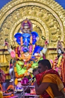 A man in traditional attire is seen speaking into a microphone in front of a large and intricately decorated statue of a deity, adorned with vibrant flowers and garlands. The backdrop is rich with gold embellishments and colorful fabric, indicating a ceremonial or religious setting.