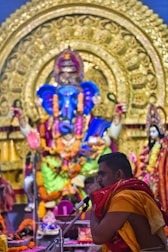 A man in traditional attire is seen speaking into a microphone in front of a large and intricately decorated statue of a deity, adorned with vibrant flowers and garlands. The backdrop is rich with gold embellishments and colorful fabric, indicating a ceremonial or religious setting.
