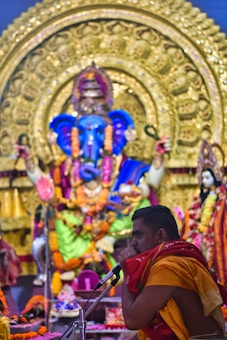 A man in traditional attire is seen speaking into a microphone in front of a large and intricately decorated statue of a deity, adorned with vibrant flowers and garlands. The backdrop is rich with gold embellishments and colorful fabric, indicating a ceremonial or religious setting.