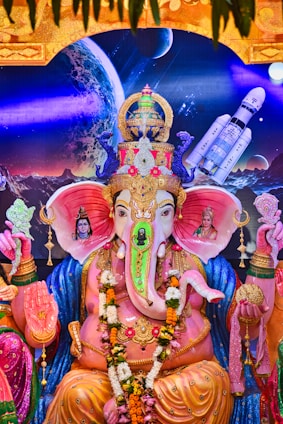 A vibrant and detailed statue of Lord Ganesha is adorned with colorful ornaments and garlands. The deity is seated in a traditional pose with a backdrop depicting cosmic elements like a planet and a spacecraft. The statue is surrounded by intricate patterns and figures of divine beings, showcasing a blend of spirituality and modern imagery.
