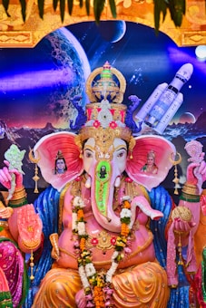 A vibrant and detailed statue of Lord Ganesha is adorned with colorful ornaments and garlands. The deity is seated in a traditional pose with a backdrop depicting cosmic elements like a planet and a spacecraft. The statue is surrounded by intricate patterns and figures of divine beings, showcasing a blend of spirituality and modern imagery.